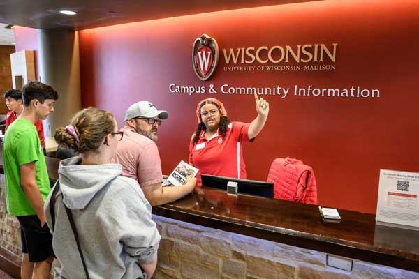 A visitor staff member behind a red 'Wisconsin Campus and Community Information' desk points directions while assisting a group of visitors.