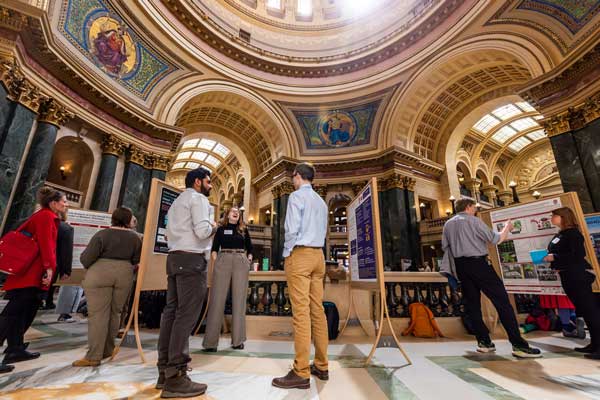 People gather around educational display boards to discuss research projects underneath the grand, ornate dome of Madison's capitol rotunda.