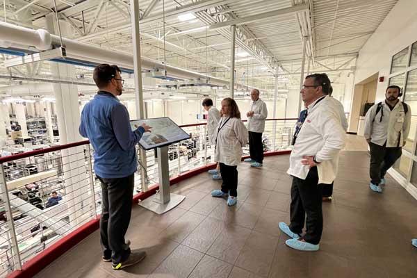 A tour guide gestures toward a digital display screen while leading a group of visitors in white lab coats along an elevated walkway overlooking a large industrial facility.
